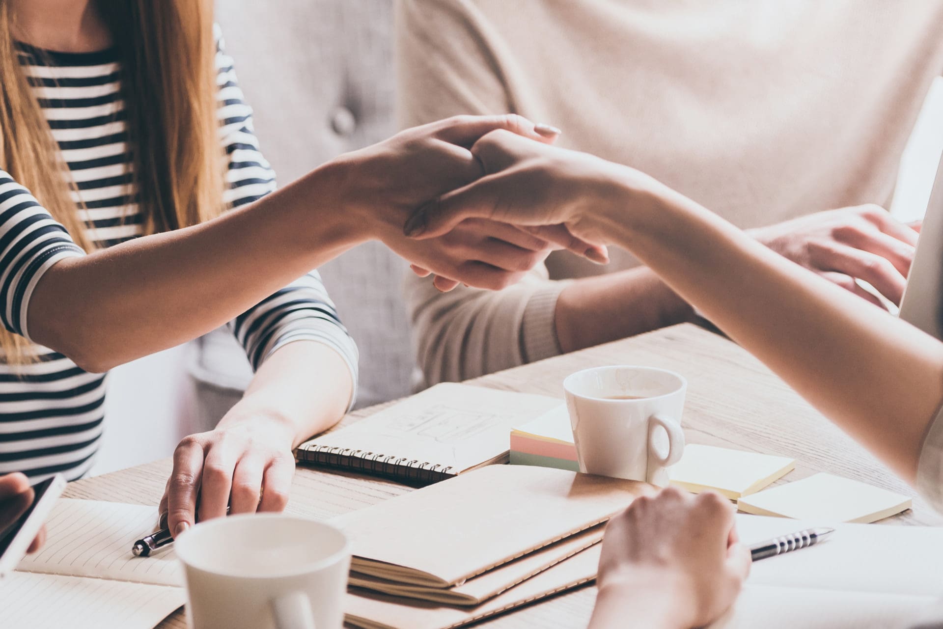 Two people shaking hands across a table with coffee cups and notes, symbolizing a successful negotiation or accepted offer.