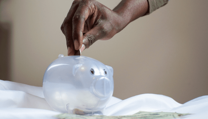 A hand putting coins into a clear plastic piggy bank to avoid being house poor.