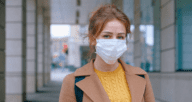 A blonde in a yellow sweater and tan jacket wearing a face mask due to coronavirus.