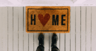 Feet standing at a welcome mat that says "Home" with a heart for the O, on a white porch
