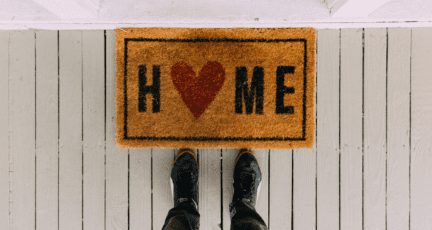 Feet standing at a welcome mat that says "Home" with a heart for the O, on a white porch