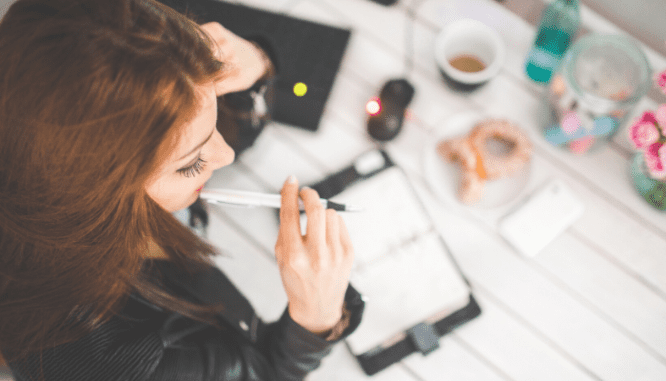 The downward view of a woman with a pen in her mouth, studying a planner in front of her.