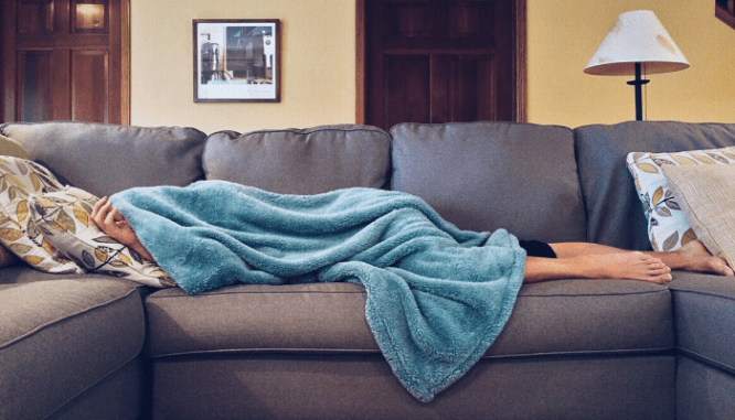 A person lying under a blue throw blanket on a gray couch with only their feet sticking out.