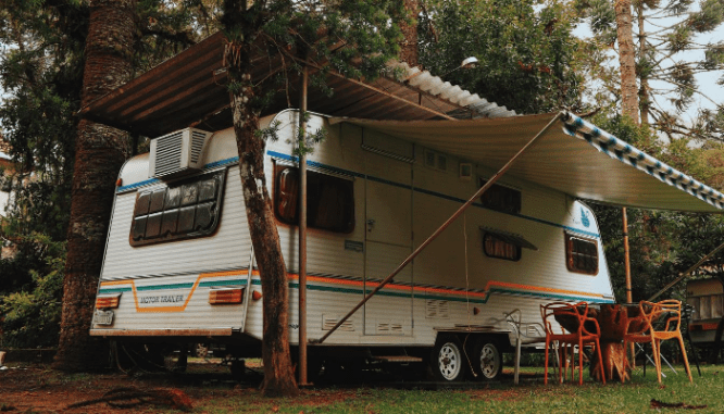 An RV parked in the forest with a patio awning set up and a table and chairs set up underneath, something you might find living off the grid.