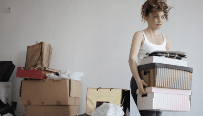 A woman with curly hair carrying moving boxes into a new house, with moving boxes stacked behind her.