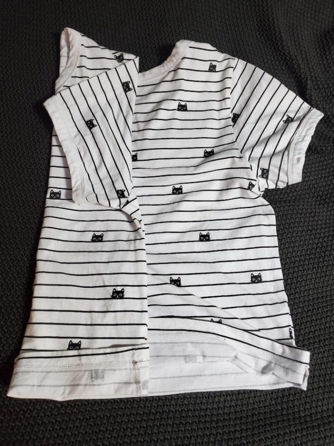 A shirt with a lined cat print with its first (left) side folded in half and the sleeve folded in for a military roll to show how to pack clothes for moving