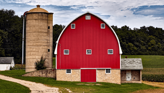 A red barn and grain silo on a green hillside with a dirt road in front.