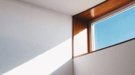 A wooden trimmed window with blue sky letting a beam of sunlight onto a white wall.
