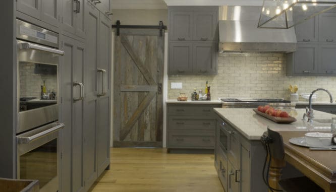 A trendy barn door that's in a kitchen with grey cabinets.