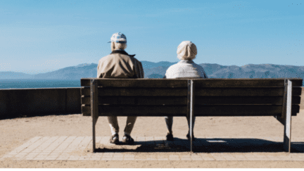An elderly couple with dementia sitting on a bench near their house.