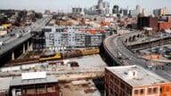 An aerial shot of downtown Kansas City showing a train on the tracks, cars on the highway, and the skyline in the distance.
