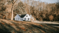 Small white farmhouse in South Carolina with black shutters and a wood barn, seen from a distance and surrounded by trees and yellowing grass.