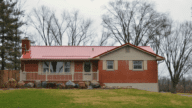 A red brick ranch-style home with a red metal roof and beige trim. There's a grassy front lawn and trees behind the house.