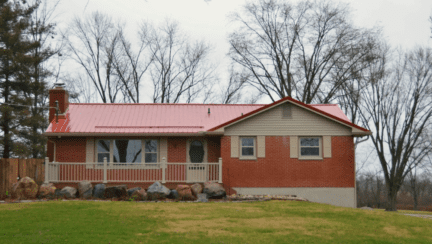 A red brick ranch-style home with a red metal roof and beige trim. There's a grassy front lawn and trees behind the house.