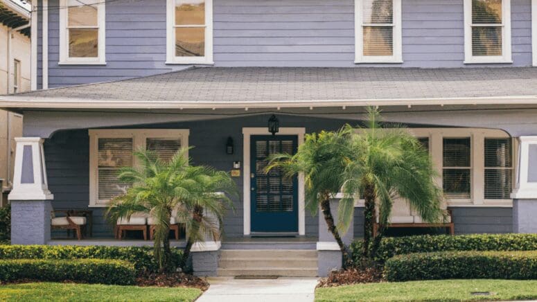 A violet craftsman-style house in Tampa with a dark blue, paned-glass front door, and small palm trees framing the entrance to the house.