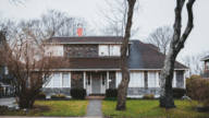 Two-story house with weathered shake siding and a sage green front door. The house has a grassy front lawn with bare trees and the street in front of it looks wet as if it had just rained.