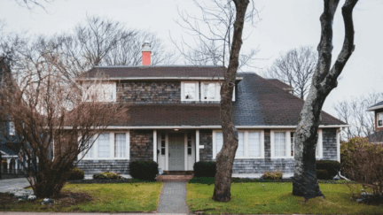 Two-story house with weathered shake siding and a sage green front door. The house has a grassy front lawn with bare trees and the street in front of it looks wet as if it had just rained.