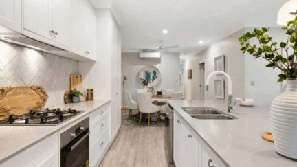 White kitchen with light gray countertops and a herringbone-patterned backsplash. The kitchen overlooks a small dining area with a round table and four upholstered chairs.