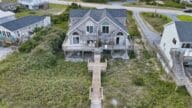 Drone shot of a gray, multi-story beach house on Topsail Beach in North Carolina. There is a wooden boardwalk that extends from the house to the sandy beach.